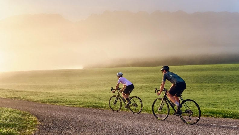 Zwei Radfahrer fahren auf einer ruhigen Landstraße durch eine grüne Hügellandschaft im warmen Morgen- oder Abendlicht.