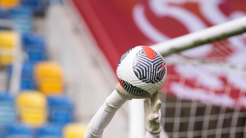 Torwarthandschuhe fangen einen Fußball vor dem Tornetz. Im Hintergrund sind unscharfe Tribünensitze zu erkennen.