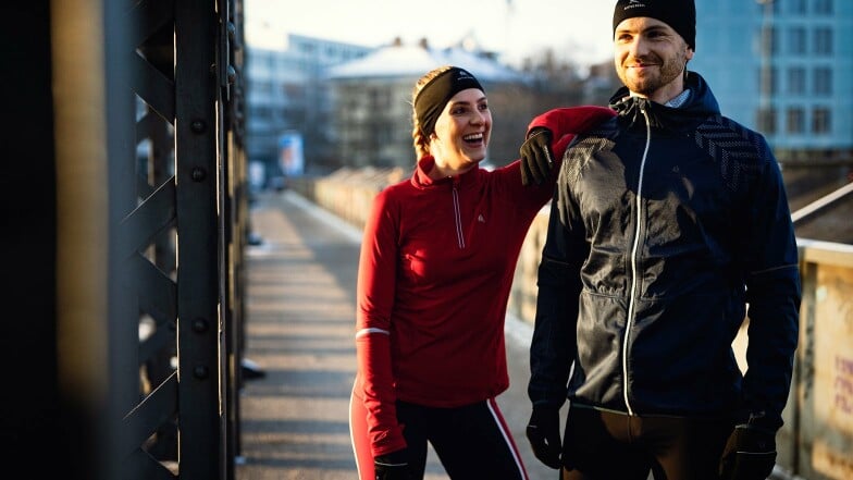 Ein sportliches Paar in Laufkleidung lacht auf einer Brücke.