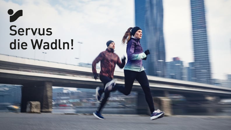 Zwei Läufer beim gemeinsamen Lauf über eine Brücke in winterlicher Stadtkulisse mit dem Text Servus die Wadln!