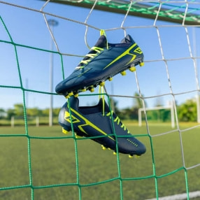 Ein Paar Fußballschuhe hängt im Tornetz auf einem Fußballplatz. Der Fokus liegt auf den Schuhen vor unscharfem Hintergrund.