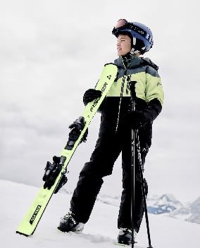 Kind mit Fischer-Ski und Skistöcken in schwarzer und hellgrüner Skibekleidung vor verschneiter Berglandschaft