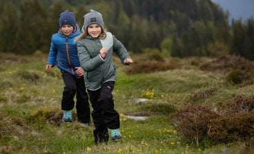 Zwei Kinder in Outdoor-Kleidung laufen lachend über eine Wiese in den Bergen.