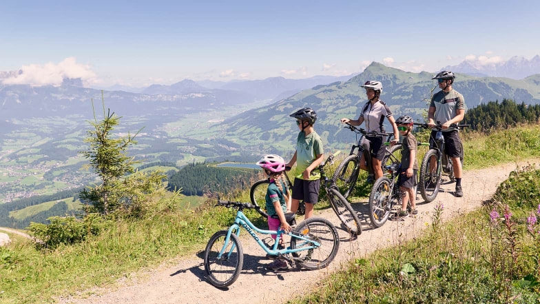 Eine Familie mit zwei Erwachsenen und drei Kindern steht mit Fahrrädern auf einem Bergweg. Sie blicken in die weite Landschaft mit Tälern, Hügeln und einem Panorama aus Bergen.
