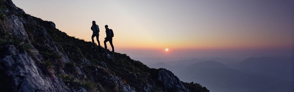 Zwei Wanderer besteigen eine Bergsilhouette und im Hintergrund ist der Sonnenuntergang zusehen.