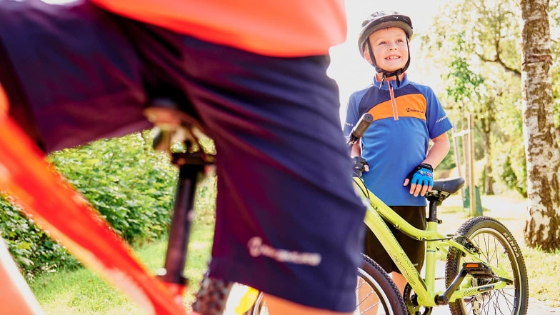 Ein Junge mit Fahrradhelm steht neben seinem Fahrrad und lacht in die Sonne. Im Vordergrund ist ein weiteres Fahrrad angeschnitten zu sehen. Die Szene spielt sich auf einem Radweg im Grünen ab.