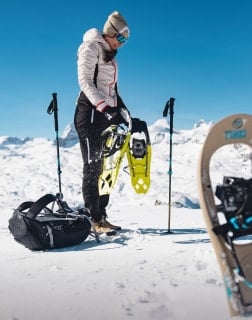 Eine Person in Winterkleidung steht im Schnee und hält gelbe Schneeschuhe, umgeben von Wanderstöcken und einem Rucksack.