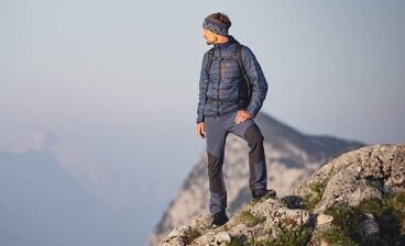 Eine Person in Wanderkleidung steht auf einem felsigen Gipfel und blickt in die Ferne vor einer Bergkulisse.