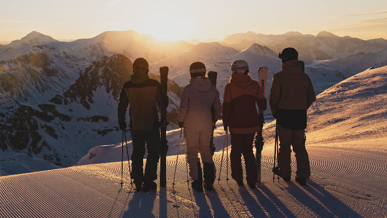 Vier Skifahrerinnen und Skifahrer stehen bei Sonnenuntergang auf einer präparierten Skipiste vor verschneiten Bergen