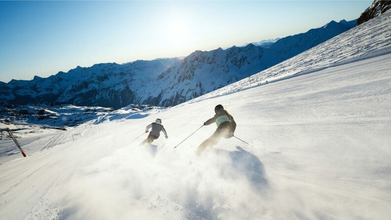 Zwei Skifahrer auf einer sonnigen Piste vor einem Bergpanorama.