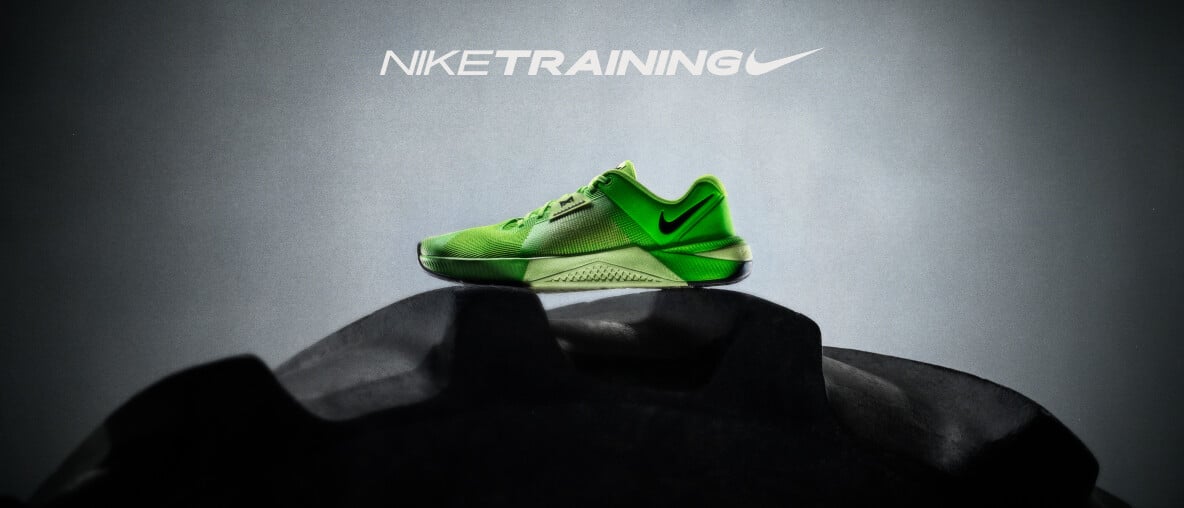 Ein grüner Nike Metcon 10 Trainingsschuh steht auf einem großen schwarzen Reifen.