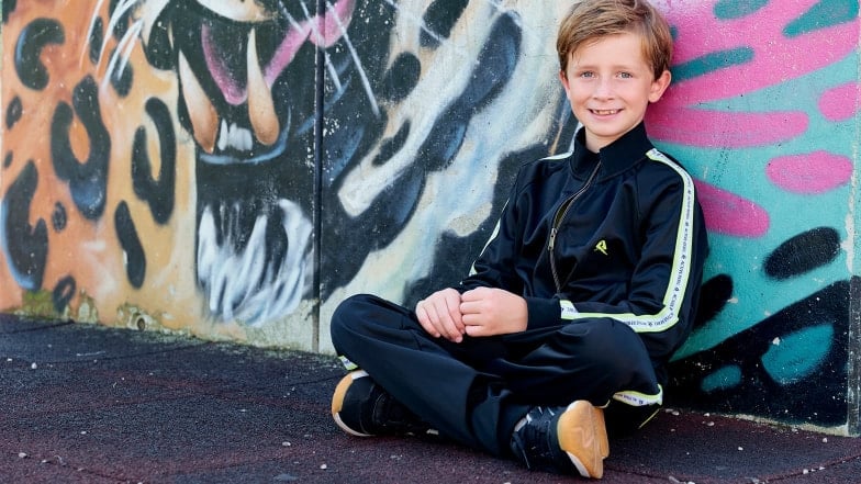 Ein Junge im Trainingsanzug sitzt lächelnd vor einer Graffiti-Wand.