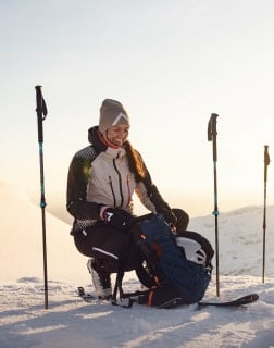 Eine Person sitzt im Schnee neben Skistöcken und einem Rucksack und lächelt in der Wintersonne.