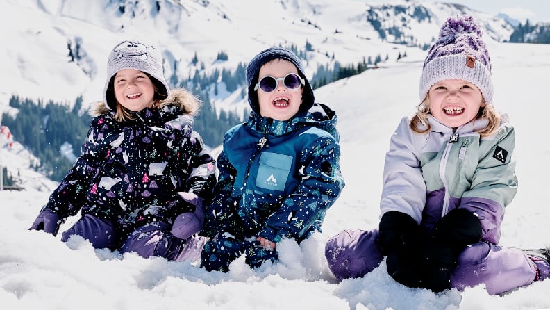 Drei lachende Kinder in Winterkleidung sitzen im Schnee vor einer verschneiten Berglandschaft