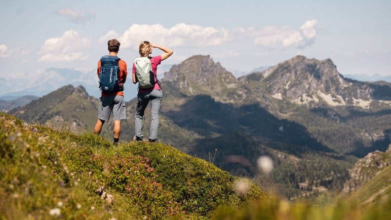 Zwei Personen mit Rucksäcken stehen auf einem Bergkamm und blicken auf eine weite Berglandschaft.