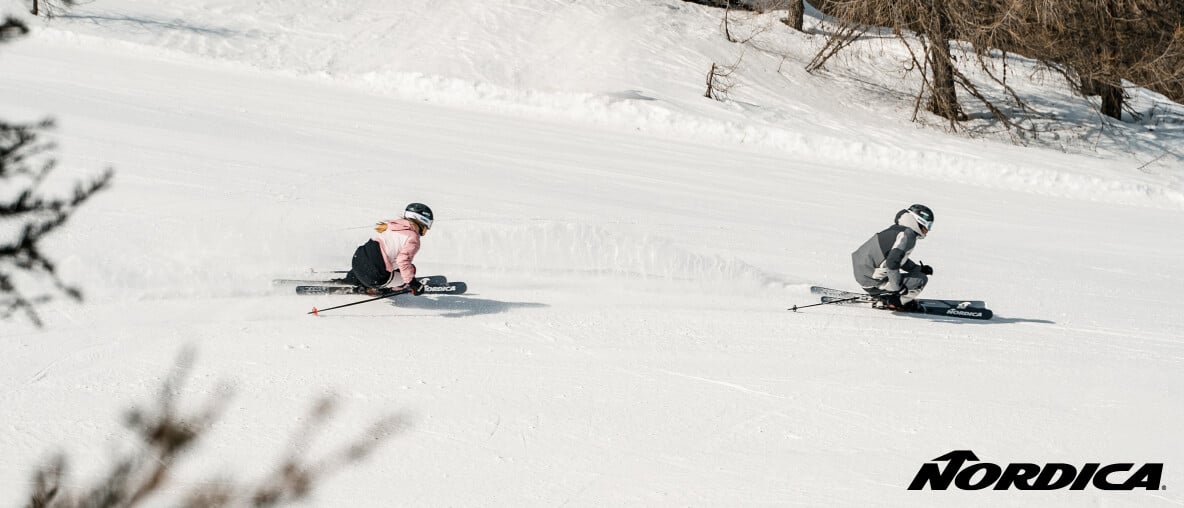 Zwei Skifahrer carven bei Sonnenschein schnell eine Piste hinunter.