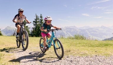 Ein Kind und eine Frau fahren gemeinsam mit dem Fahrrad über einen Schotterweg auf einer Alm, im Hintergrund sind Berge und Wälder zu sehen. Beide tragen Fahrradhelme und Sportbekleidung.