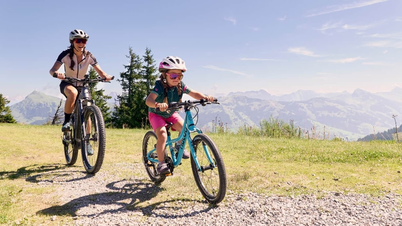 Ein Kind und eine Frau fahren gemeinsam mit dem Fahrrad über einen Schotterweg auf einer Alm, im Hintergrund sind Berge und Wälder zu sehen. Beide tragen Fahrradhelme und Sportbekleidung.