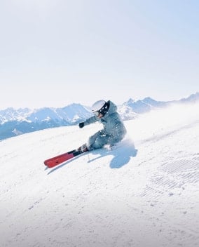 Eine Person fährt mit Skiern eine verschneite Piste hinunter, umgeben von Berggipfeln.