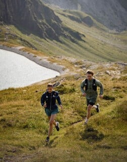 Zwei Personen laufen in Sportkleidung über eine grüne Bergwiese neben einem See.
