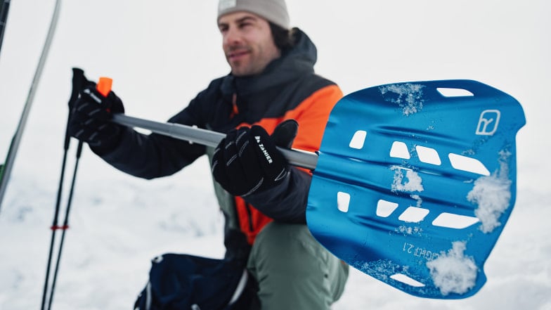 Ein Mann in Winterausrüstung hält eine blaue Lawinenschaufel im Schnee.