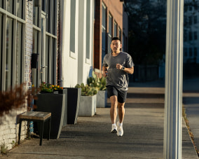 Ein Mann joggt auf einem Bürgersteig entlang einer Reihe von Gebäuden. Er trägt ein graues T-Shirt, schwarze Shorts und weiße Laufschuhe. Die Sonne scheint von links, wodurch der Bürgersteig und die Gebäudefassade beleuchtet sind. Im Hintergrund sind Pflanzen in großen Töpfen vor den Gebäuden zu sehen. Der Mann blickt nach vorne und trägt eine Smartwatch am linken Handgelenk.