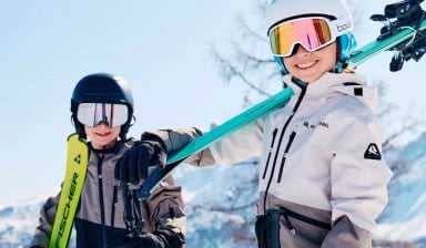 Dva mladí lyžaři s helmami a lyžemi na ramenou, úsměv na tváři a jasné počasí v horách.