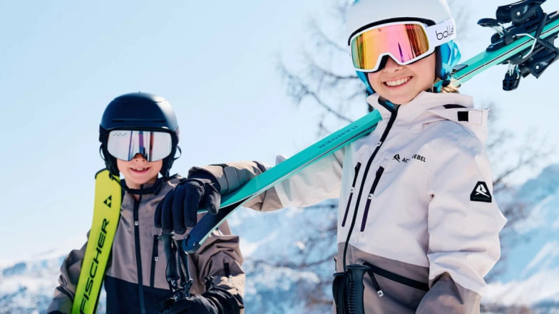 Dva mladí lyžaři s helmami a lyžemi na ramenou, úsměv na tváři a jasné počasí v horách.