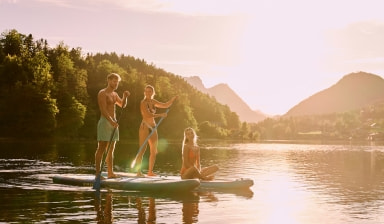 Drei Personen mit zwei SUPs auf einem See bei untergehender Sonne.