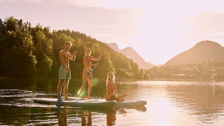 Drei Personen mit zwei SUPs auf einem See bei untergehender Sonne.