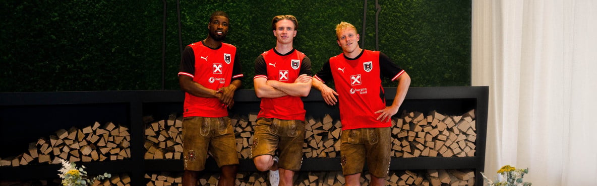 Drei Fußballspieler in roten ÖFB-Trikots und ÖFB-Lederhosen stehen vor gestapeltem Brennholz.