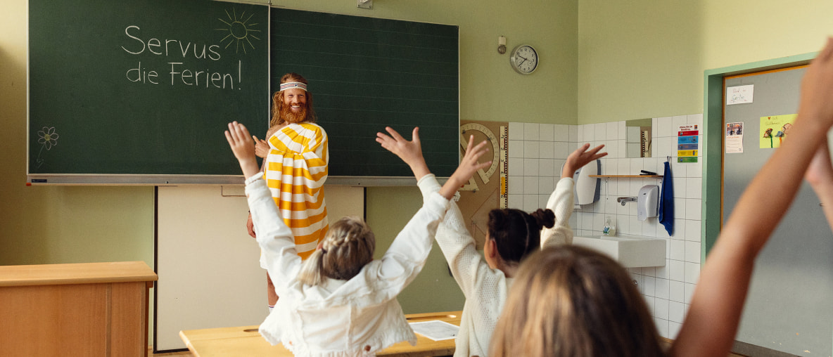 Der Sommer (Mann mit Bart und luftigem, gelbgestreiftem Umhang) steht in einer Schulklasse und zeigt auf die Tafel, auf der "Servus, die Ferien!" steht. Die Kinder jubeln.