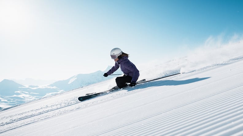 Skifahrerin mit Helm und Skibrille fährt eine präparierte Skipiste vor verschneiter Berglandschaft hinunter