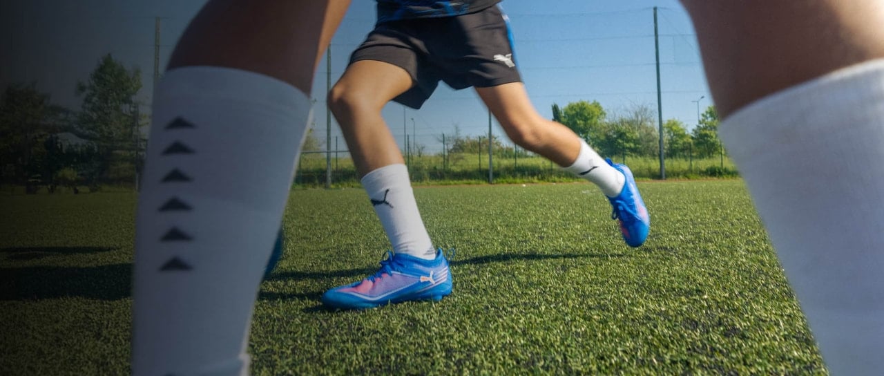 Beine von Fußballspielern mit Stutzen und Stollenschuhen auf einem Kunstrasenplatz in Bewegung. Der Fokus liegt auf den Füßen während einer Spielsituation.