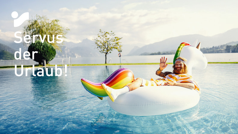 Der Sommer (Mann mit Bart und luftigem, gelbgestreiftem Umhang) entspannt auf einem aufblasbaren Einhorn im Pool mit Bergblick.