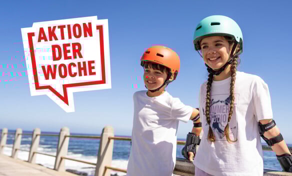 Zwei Kinder mit Helmen und Schutzausrüstung lächeln am Meer.