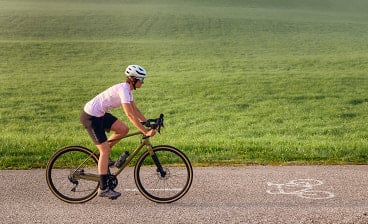 Frau fährt auf einem olivgrünen Gravel Bike auf einer asphaltierten Straße entlang einer grünen Wiese