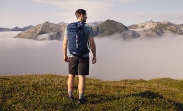 Mann mit Rucksack steht auf einer Bergwiese und blickt auf eine Berglandschaft über den Wolken.