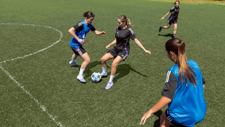 Mehrere Fußballspielerinnen stehen auf einem Kunstrasenplatz im Zweikampf um den Ball. Zu sehen sind Spielerinnen in Trainingskleidung während einer Spielsituation.