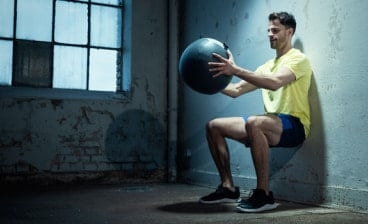 Muž cvičí v tělocvičně ve dřepu a v rukou má nafukovací fitness míč.
