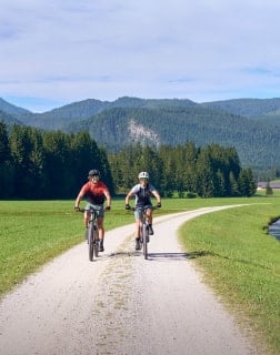 Zwei Erwachsene fahren nebeneinander auf einem Schotterweg durch eine weite Wiesenlandschaft mit Bergen.