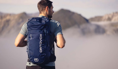 Ein Mann steht am Gipfel eines Berges und schaut ins Tal, das unter ihm im Nebel liegt. Er trägt am Rücken einen dunkelblauen Rucksack von Ortovox.