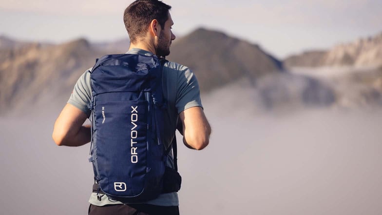 Ein Mann steht am Gipfel eines Berges und schaut ins Tal, das unter ihm im Nebel liegt. Er trägt am Rücken einen dunkelblauen Rucksack von Ortovox.