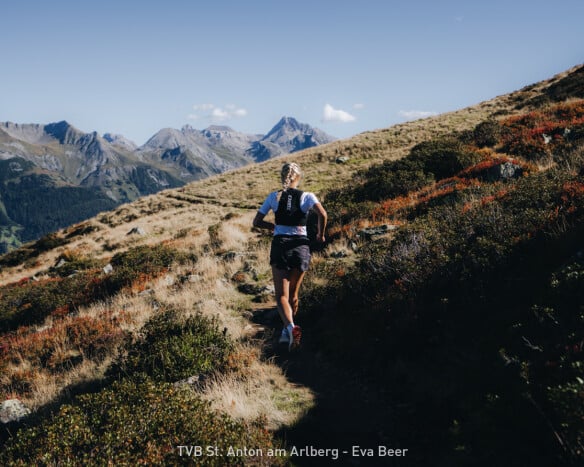 Trailrunnerin läuft auf einem alpinen Bergpfad mit Blick auf ein beeindruckendes Bergpanorama bei Sonnenschein.