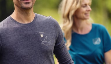 Nahaufnahme eines Mannes im grauen McKinley-Shirt; im Hintergrund ist eine Frau in einem blauen Shirt in der Natur zu sehen.