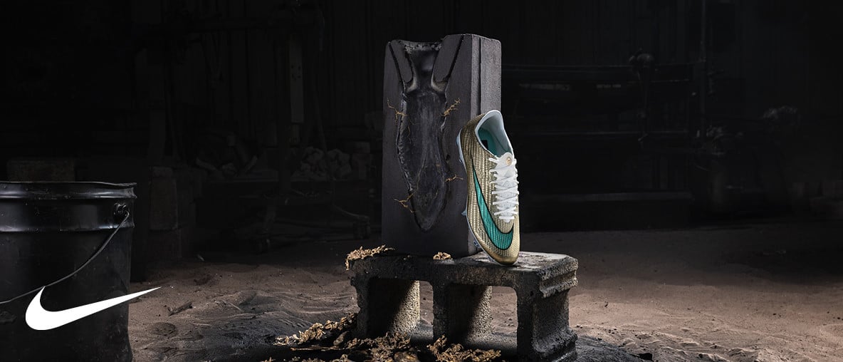 Goldfarbener Nike-Fußballschuh steht auf einer dunklen Werkbank vor einer schwarzen Metallplatte in einer Werkstatt.