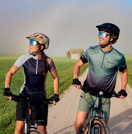 Zwei Personen mit Helmen und Fahrrädern stehen auf einem Schotterweg zwischen grünen Feldern.