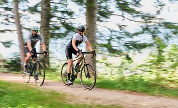 Zwei Radfahrer fahren mit Gravel Bikes auf einem Waldweg zwischen Bäumen, aufgenommen mit Bewegungsunschärfe