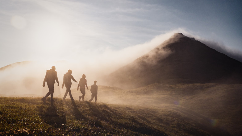 Eine Gruppe von vier Personen beim Wandern in den Bergen bei Sonnenaufgang. Nebelschwaden ziehen über einen Gipfel in der Ferne.