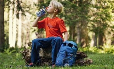 Chlapec sedí v lese s outdoorovým batohem a pije z láhve.